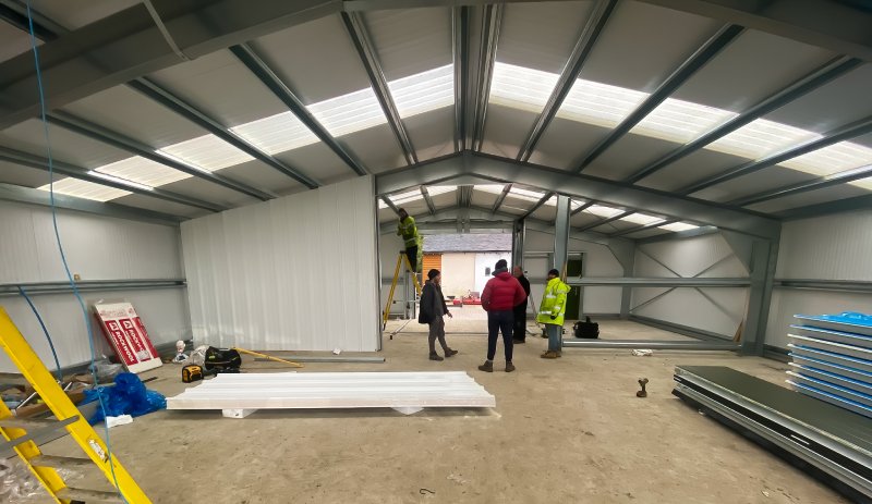 Interior view of a large steel building under construction with workers discussing the project near a central support beam and skylights.