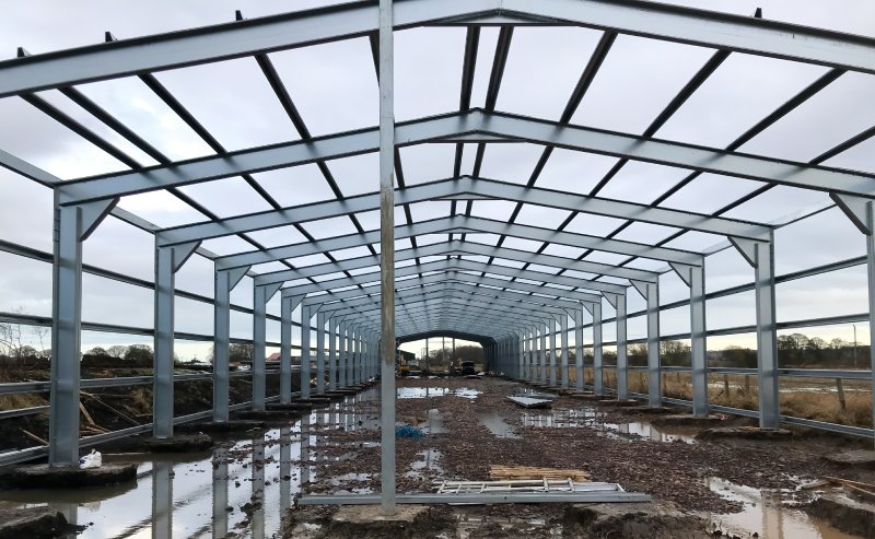 Interior view of a large steel building under construction, showing the skeletal grey metal framework, rafters, and support columns on a muddy site.