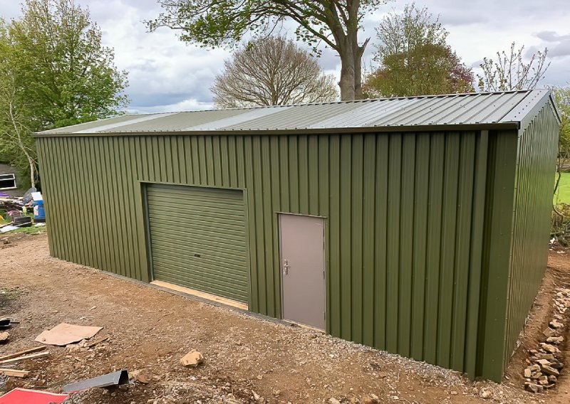 Elevated view of a green steel shed featuring translucent roof panels, a roller door, and a personnel door, surrounded by a dirt construction area.