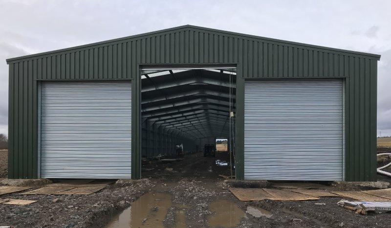A symmetrical front view of a large olive green steel warehouse with two closed silver roller doors and one open central bay showing the interior frame.